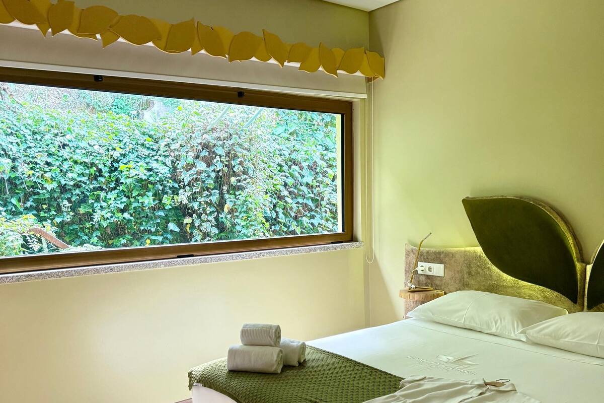 A bedroom with a large window, a bed with a green blanket, and a pair of shoes on the floor.