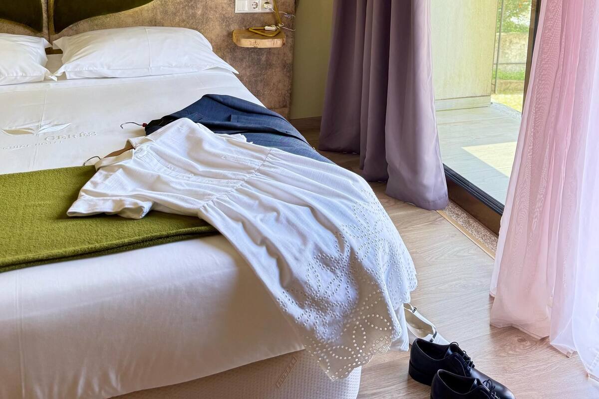 A hotel room with a bed, a bedside table with a lamp, a window with curtains, and a view of greenery outside.