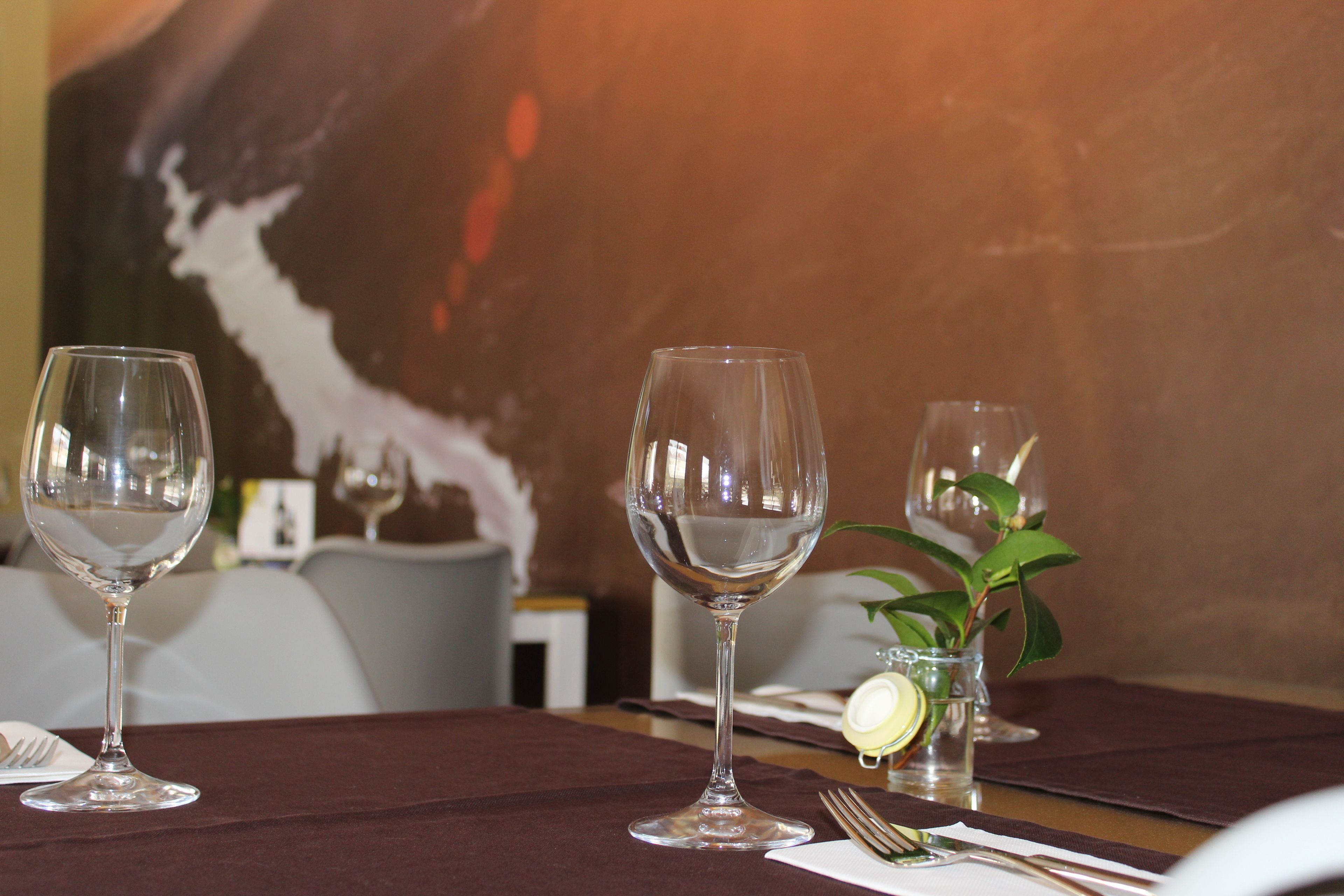 Una mesa de comedor con tres copas de vino vacías, un pequeño florero con una planta y un gajo de limón sobre un mantel.