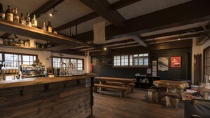 A rustic bar area with wooden shelves, a wooden counter, and a menu board. There are wooden benches and tables, and a large window providing natural light.