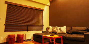 Un salón con sofá, dos taburetes de madera, una mesita de madera y un ventanal con persianas.