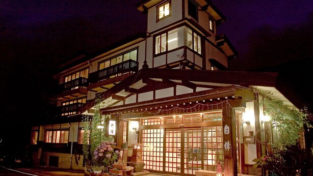 A traditional Japanese-style building with a two-story structure, wooden balconies, and a covered entrance area with potted plants and string lights.