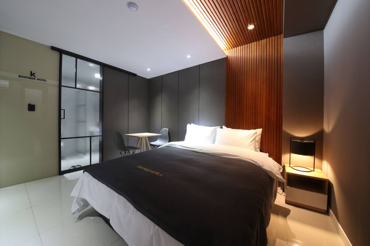 A modern hotel room with a large bed, a small desk, and a glass partition wall.