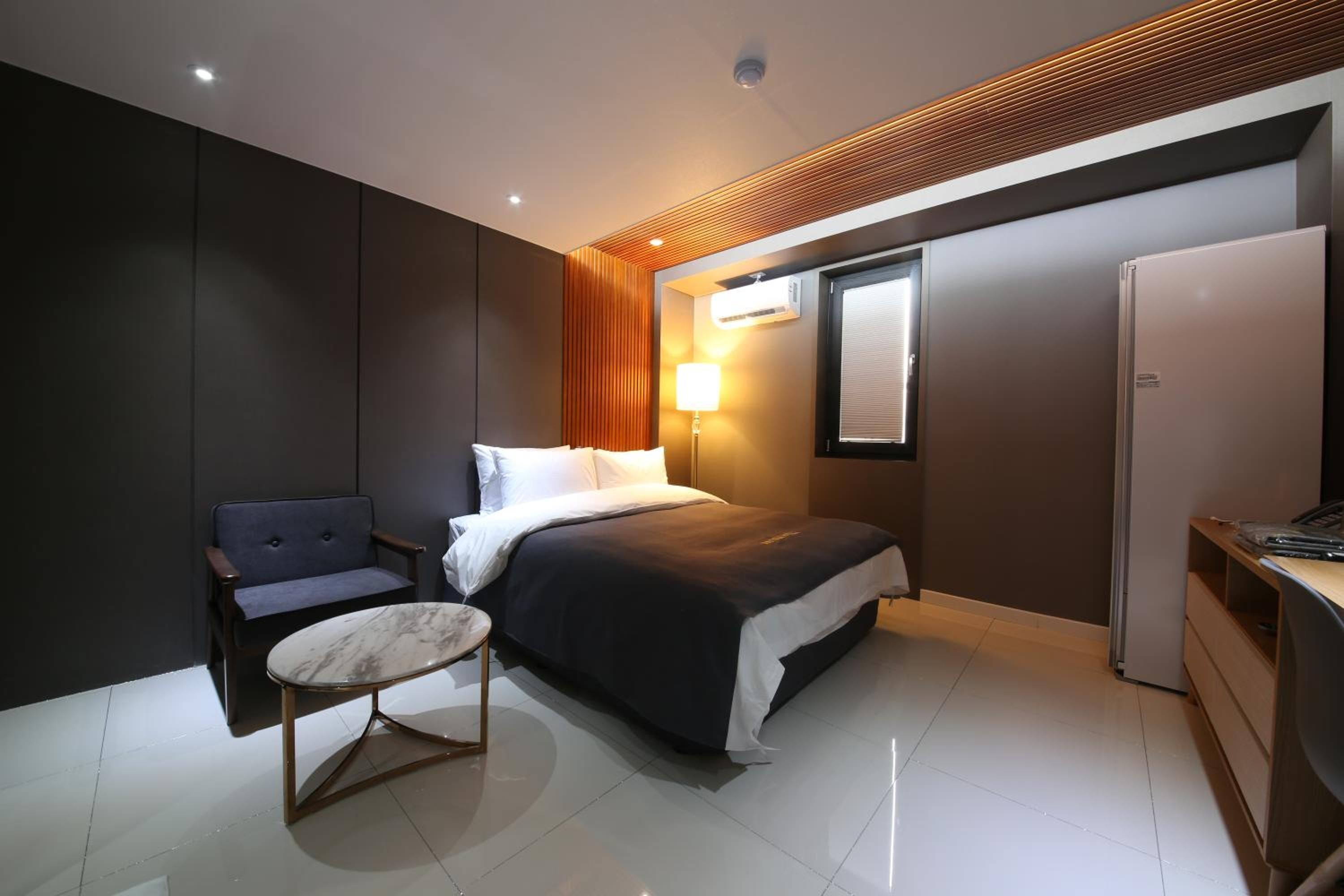 A modern hotel room with a bed, a chair, a small table, a desk, and a refrigerator.