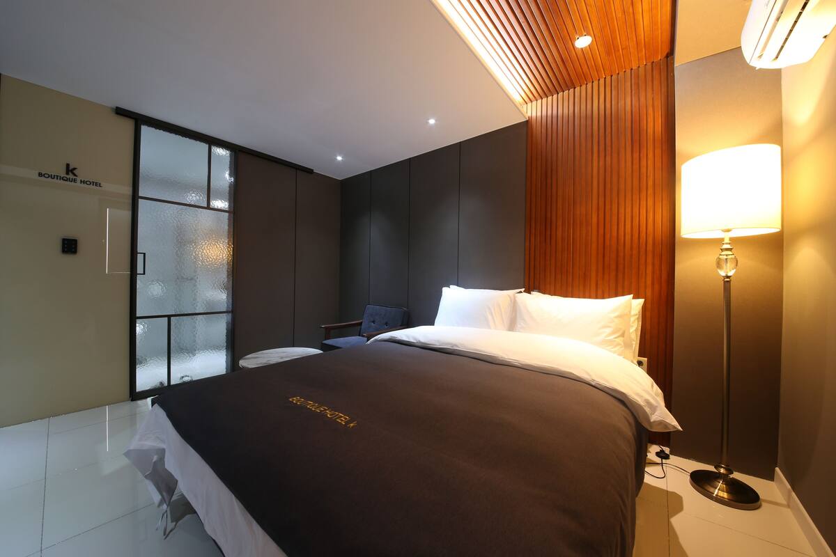 A modern hotel room with a large bed, a desk, a chair, and a window with frosted glass.
