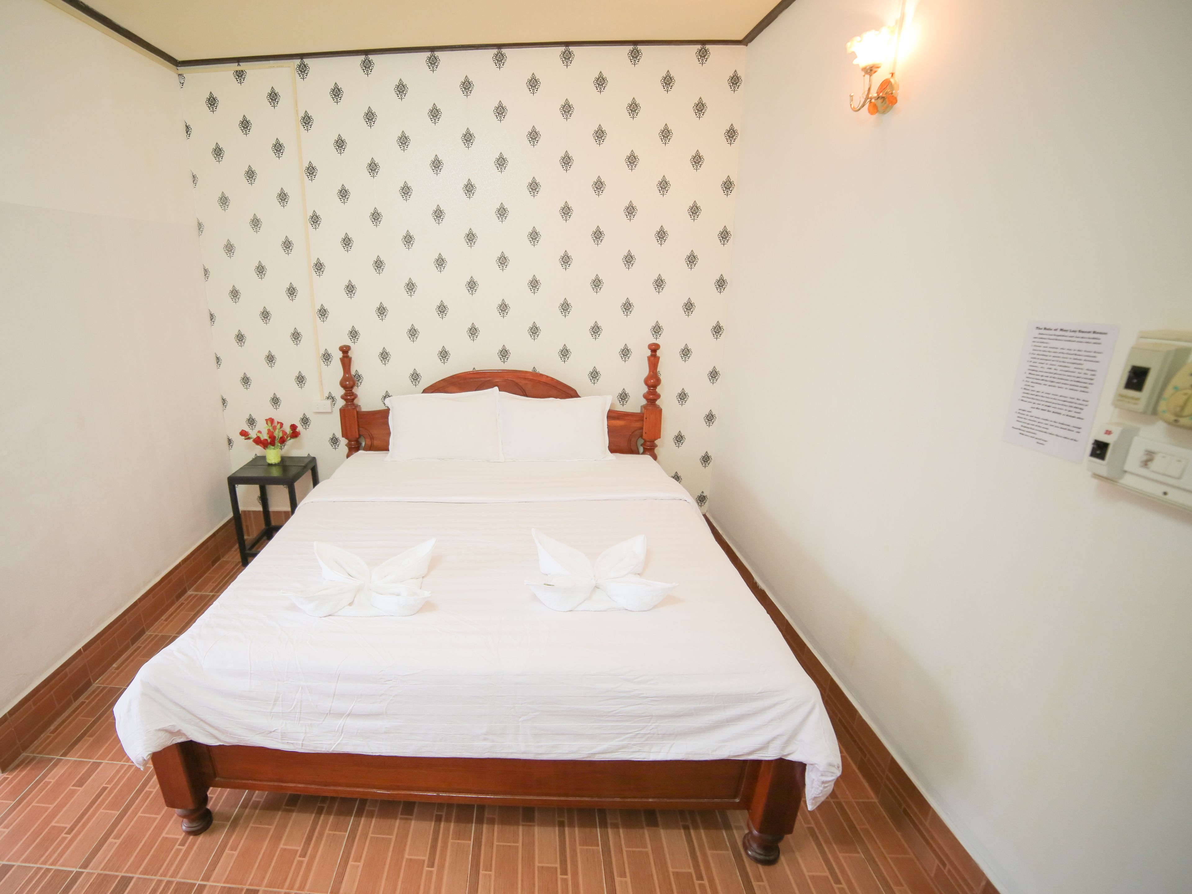 Una cama bien hecha con cabecera de madera y ropa de cama blanca, una mesita de noche con un jarrón de flores y una lámpara fijada a la pared.