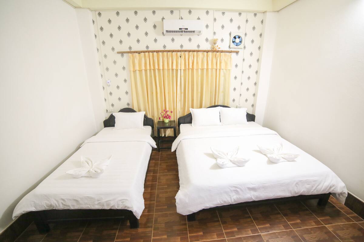 A hotel room with two single beds, a small table with a vase of flowers, and an air conditioning unit on the wall.