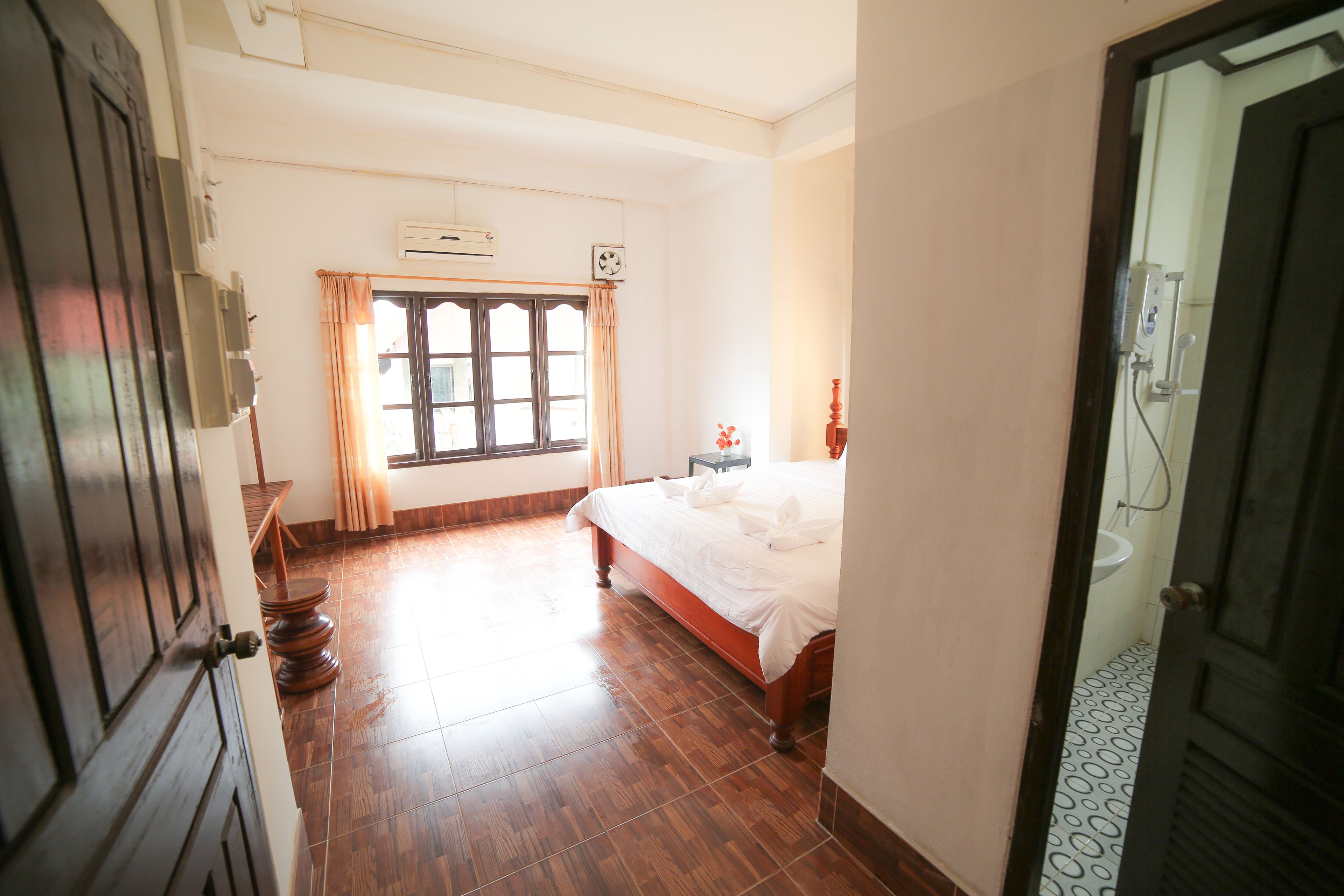 A bedroom with a wooden floor, a bed with white linens, a window with curtains, and a bathroom with a sink and shower.