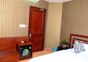 A hotel room with a wooden door, a small bedside table with a phone and a remote, a nightstand with a vase of flowers, and a bed with a colorful pillow.