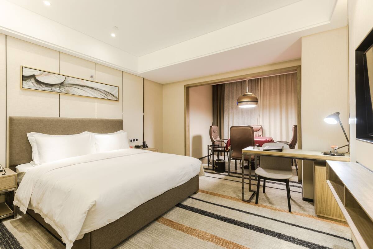 A hotel room with a large bed, a desk, and a chair. There is a painting on the wall and a window with curtains.