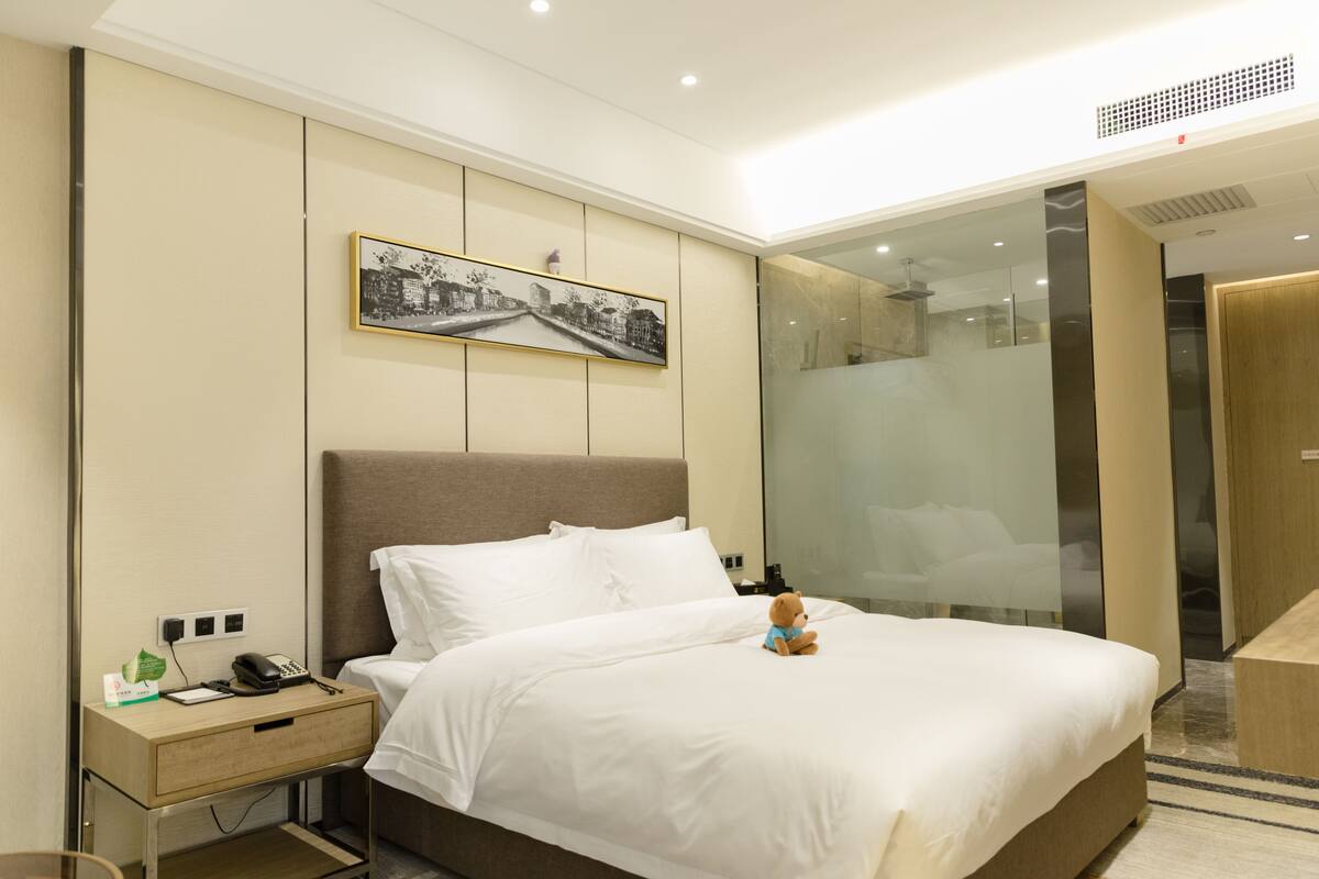 A hotel room with a large bed, a nightstand with a phone, a framed picture on the wall, and a glass shower enclosure.