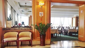 Un hall de hotel con una recepción, dos sillones, una planta en maceta, una araña y una zona de comedor al fondo.