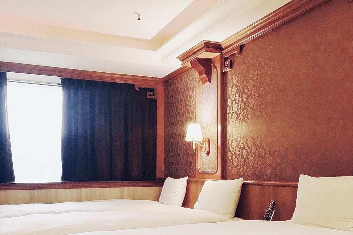 Ein Hotelzimmer mit zwei Betten, einem großen Fenster mit Vorhängen, einer Wandlampe und einem Holz-Kopfteil.