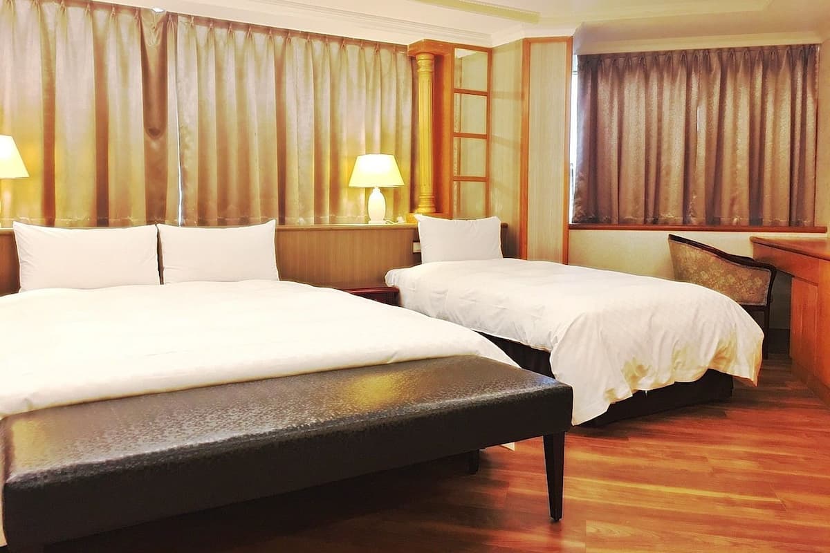 Ein Hotelzimmer mit zwei Betten, einer hölzernen Bank, einem Nachttisch mit Lampe und einem großen Fenster mit Vorhängen.