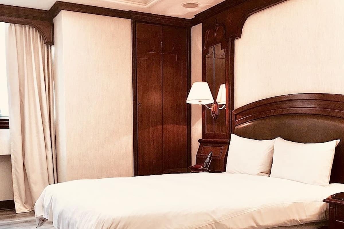 Ein großes Bett mit Holz-Kopfteil, ein Nachttisch mit Lampe und ein großes Fenster mit Vorhängen.