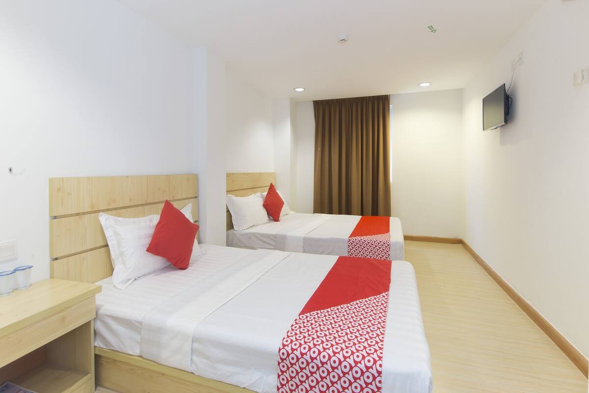 Ein Hotelzimmer mit zwei Betten, jedes mit weißer Bettwäsche und roten Akzenten, einem hölzernen Kopfteil, einem Nachttisch mit Lampe, einem an der Wand montierten Fernseher und einem Fenster mit Vorhängen.
