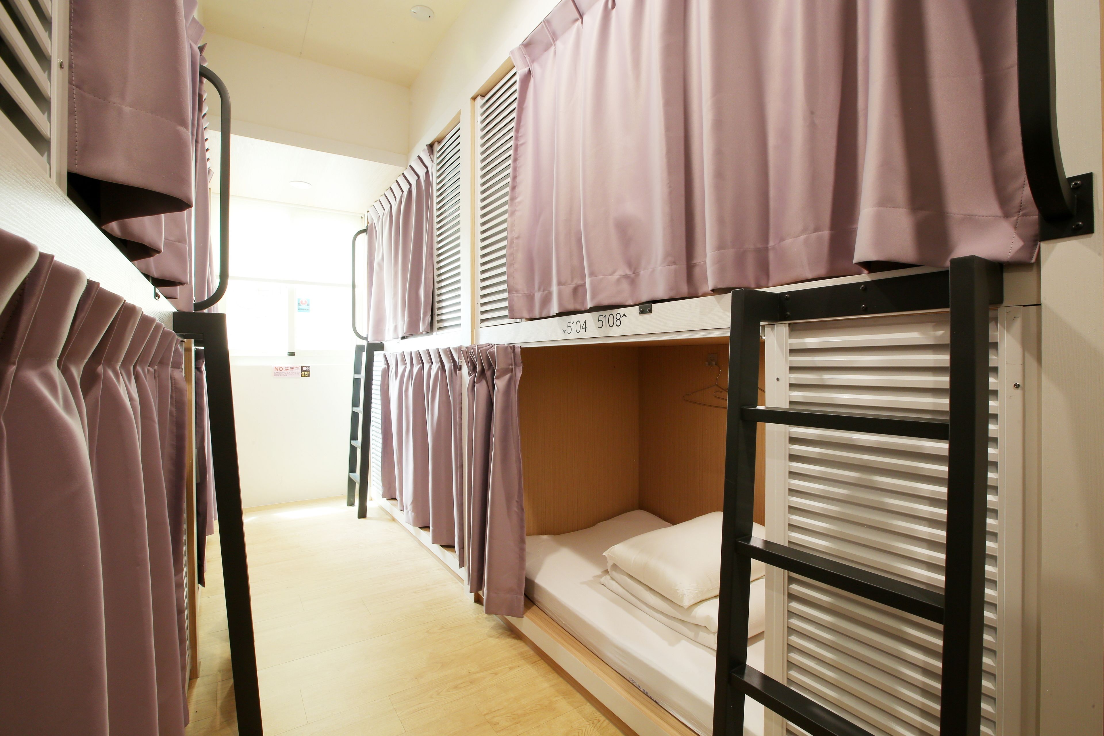 A dormitory room with bunk beds, a wooden floor, and a window with blinds.