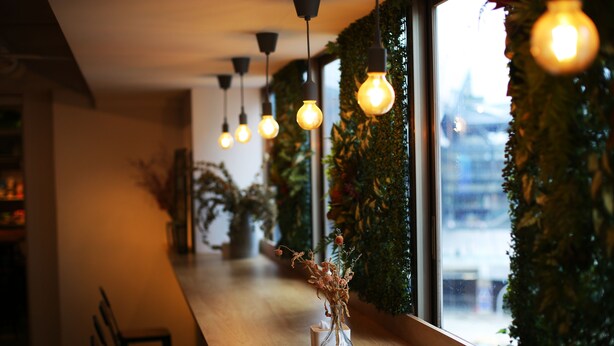 A restaurant interior with a long wooden counter, hanging light bulbs, and a vase with dried flowers. - Loosha Hostel (Taichung)
