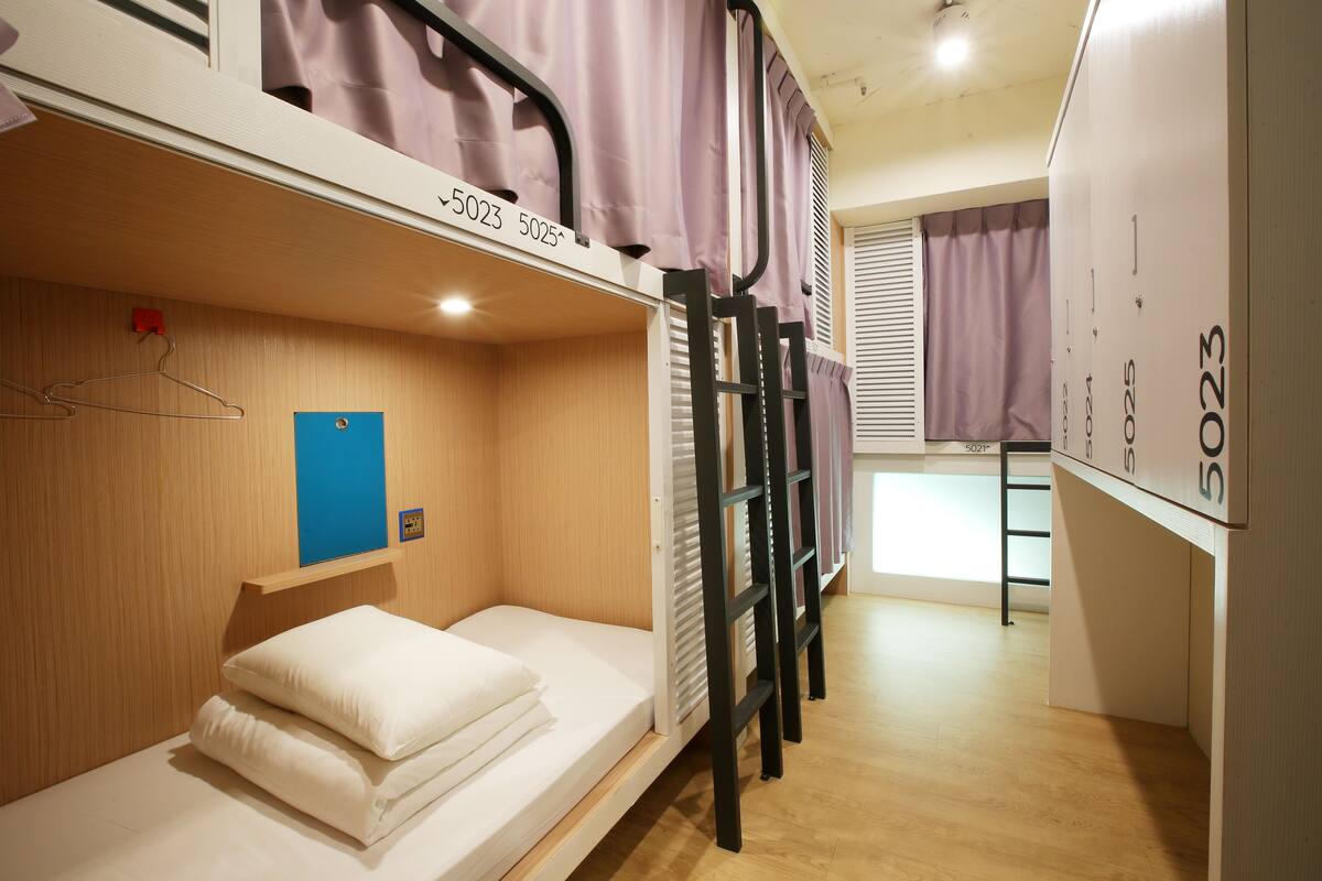 A dormitory room with bunk beds, a wooden floor, a blue door, and a window with curtains.