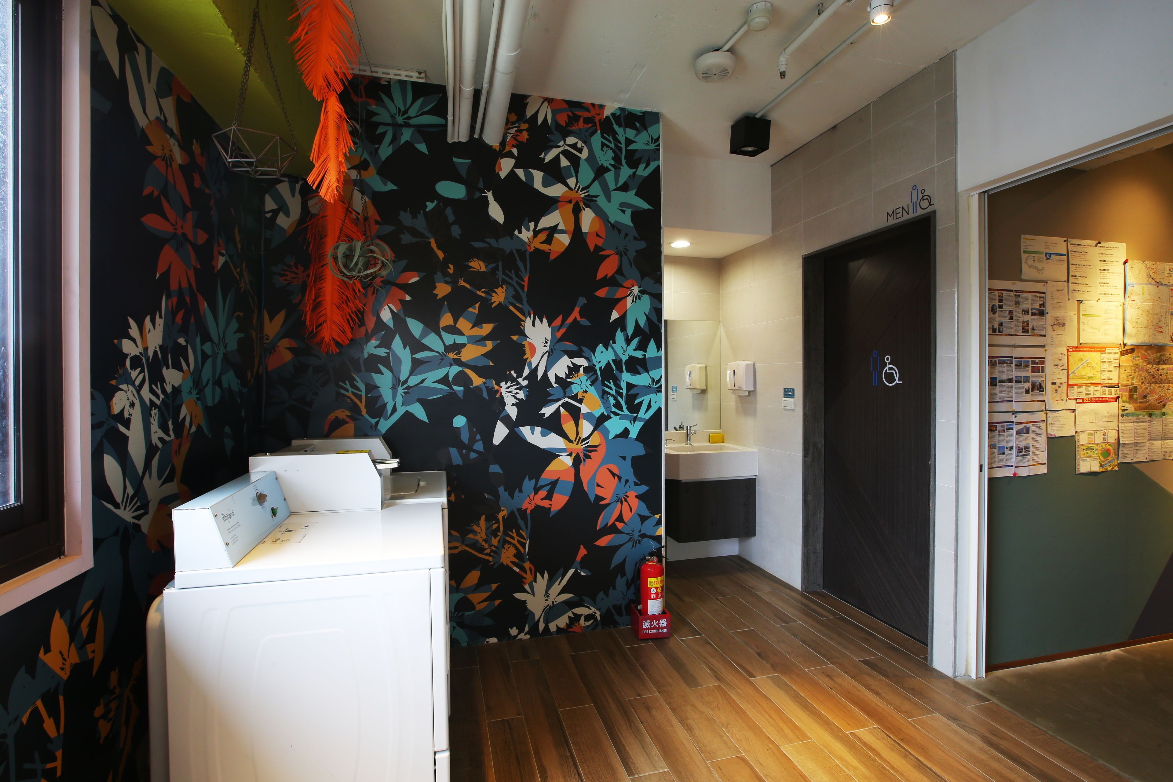 Un vestíbulo de hotel con suelo de madera, un mostrador de recepción blanco, un muro con motivos florales de colores, un extintor y un letrero para el baño de hombres.