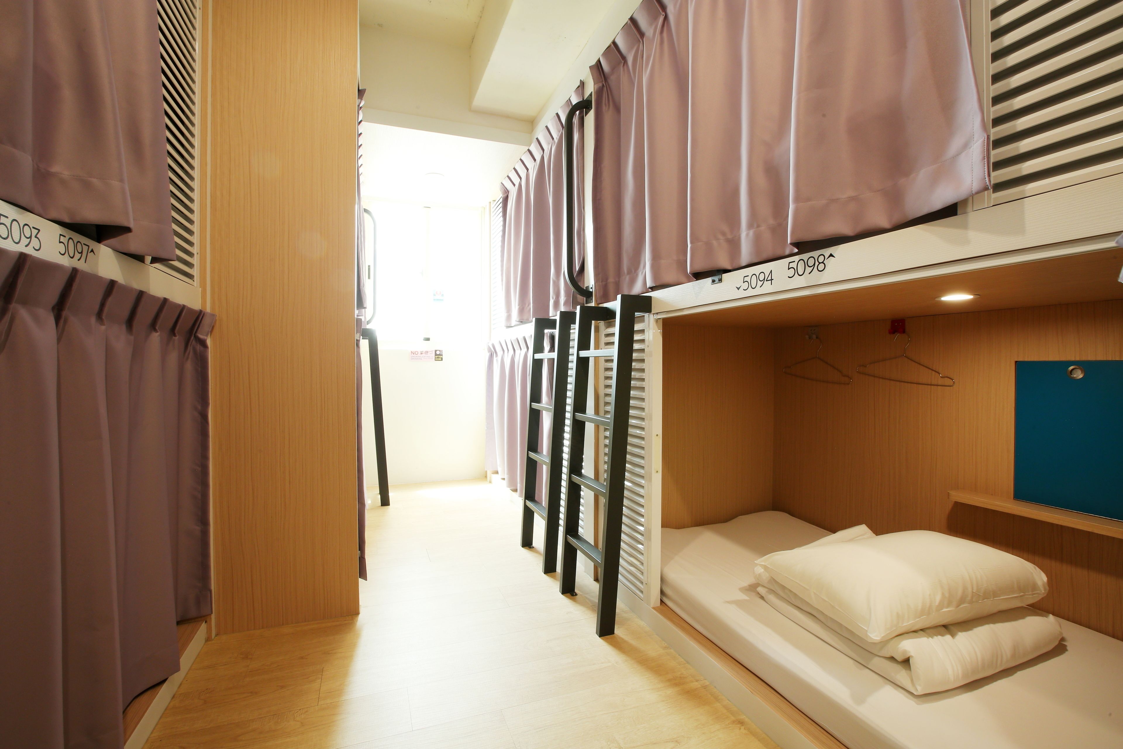 A dormitory room with bunk beds, a wooden floor, and a window with blinds.