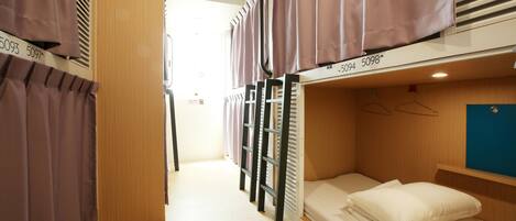 A dormitory room with bunk beds, a wooden floor, and a window with blinds.