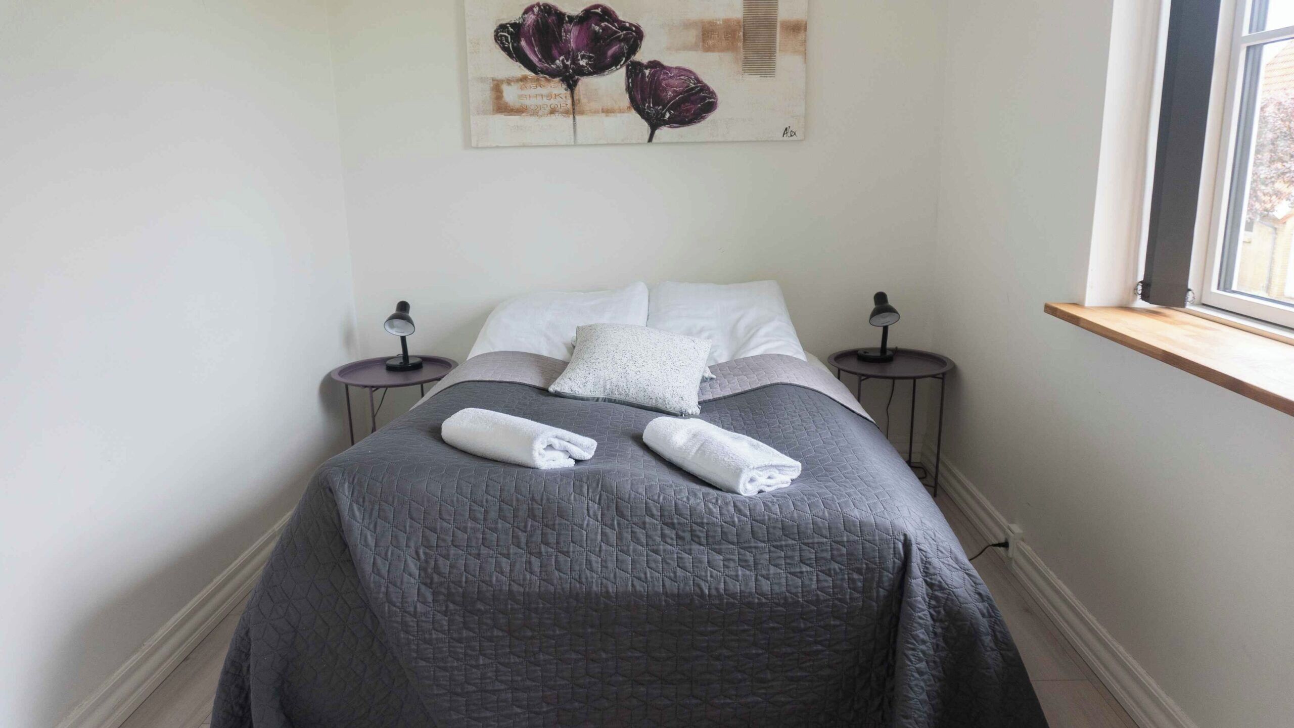 A neatly made bed with a gray quilt, two white pillows, and two rolled towels. There are two bedside tables with lamps, and a painting of flowers on the wall.