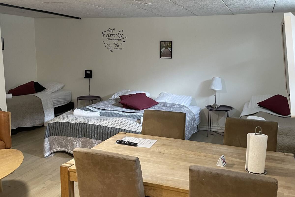 A compact, well-lit room with a bed, a small table with chairs, a bedside table with a lamp, and a wall decoration.