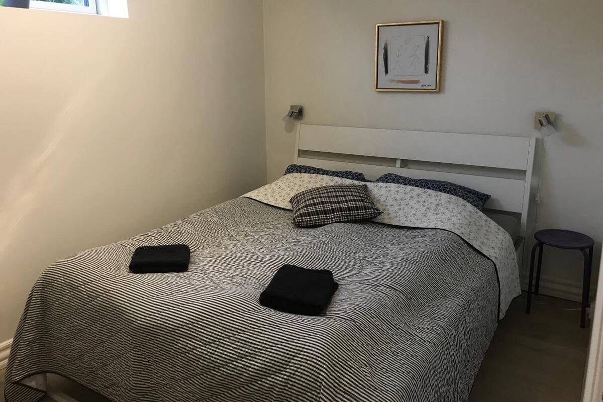 A neatly made bed with a patterned bedspread and pillows, a small stool, and a framed picture on the wall.