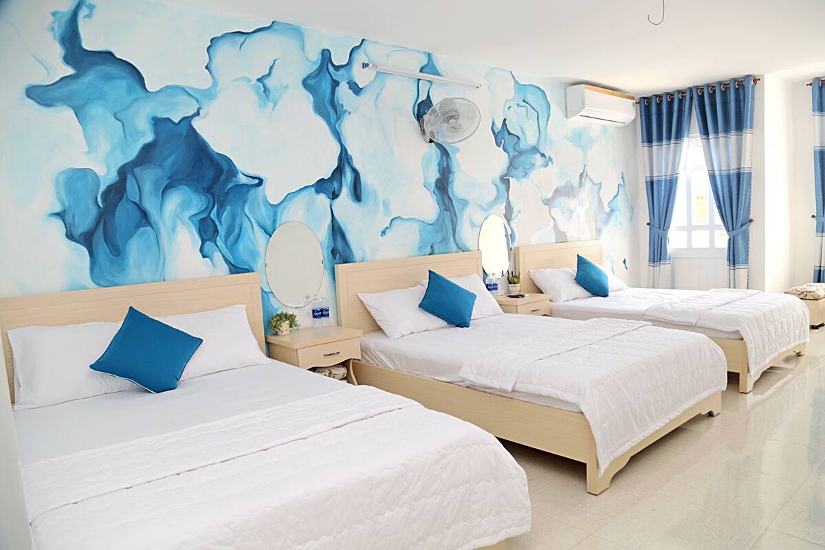 A hotel room with three beds, a large abstract mural on the wall, a ceiling fan, and a window with curtains.
