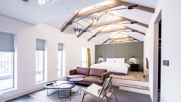 A modern hotel room with a loft-style ceiling, a large bed, a sofa, a small table, and a chair. - Patio7 Hotel (Seoul)