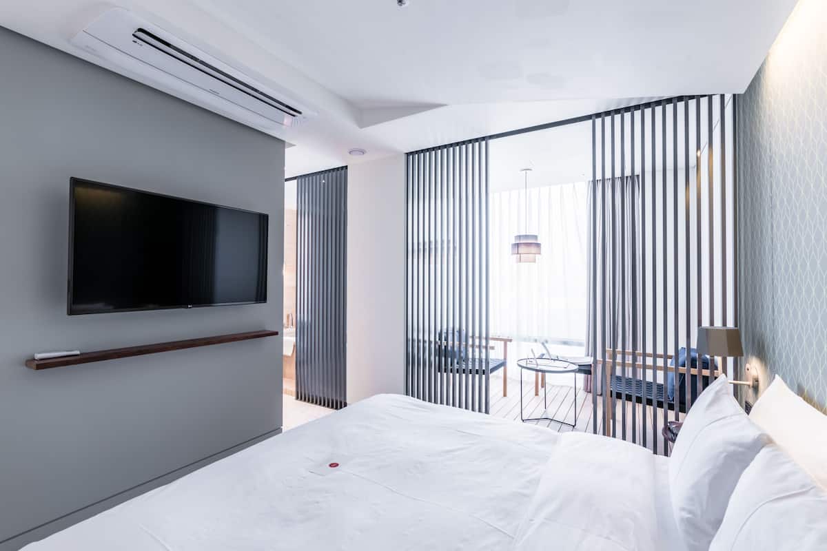 Habitación de hotel moderna con cama, televisor montado, una mesita pequeña y un balcón con vistas.