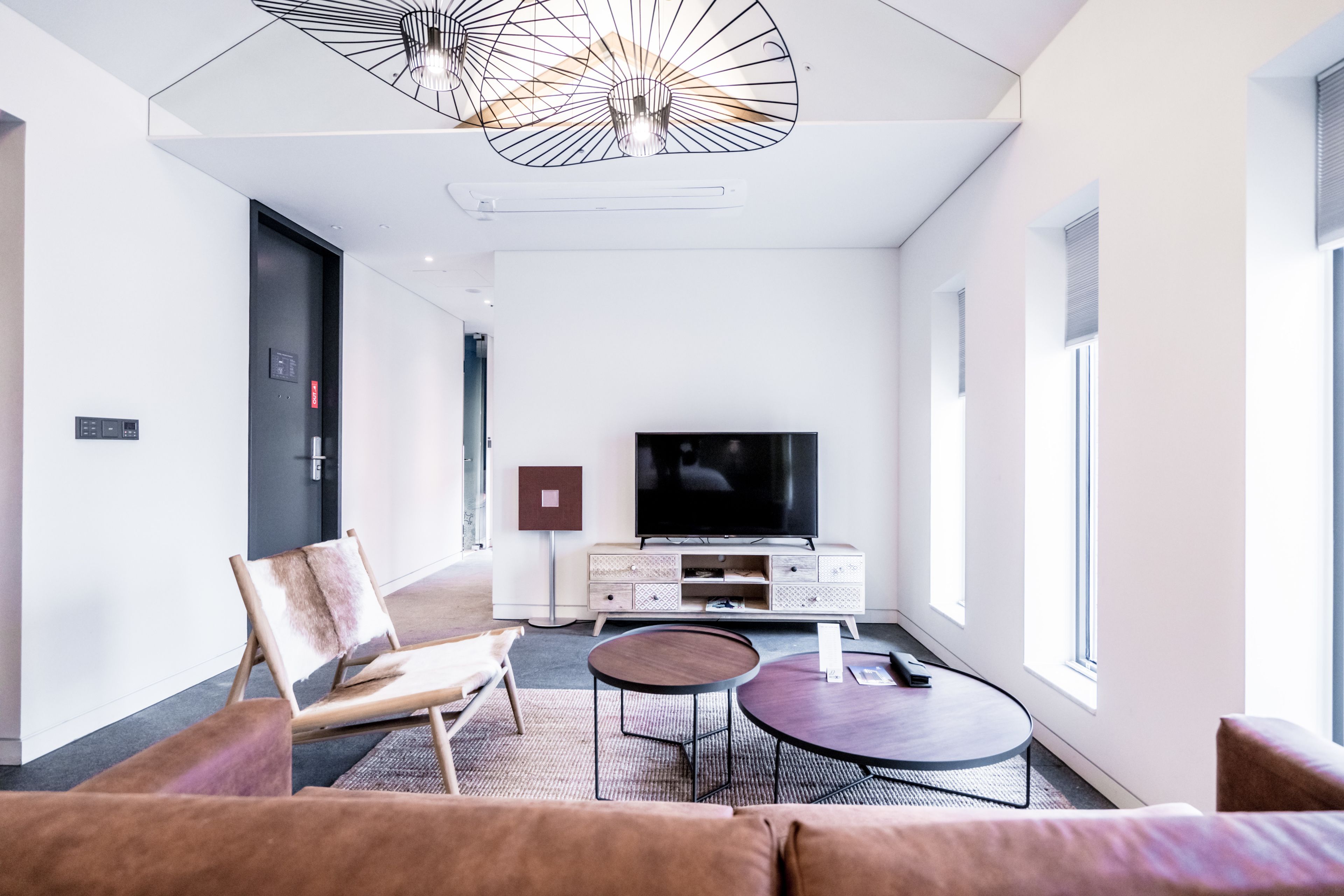 A modern hotel room with a sofa, a round coffee table, a TV, and a ceiling light fixture.