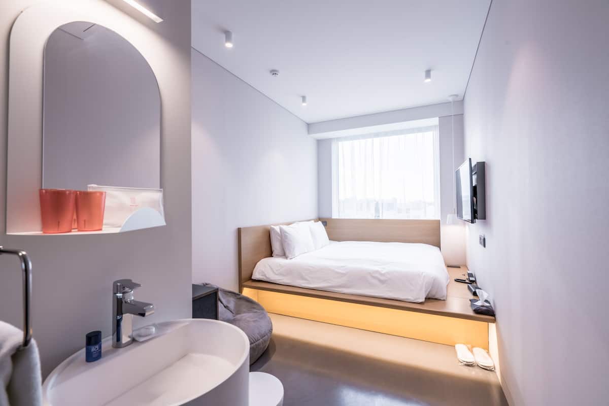 Una habitación de hotel moderna con una cama grande, una banca empotrada, un espejo amplio, una bañera y un televisor montado en la pared.