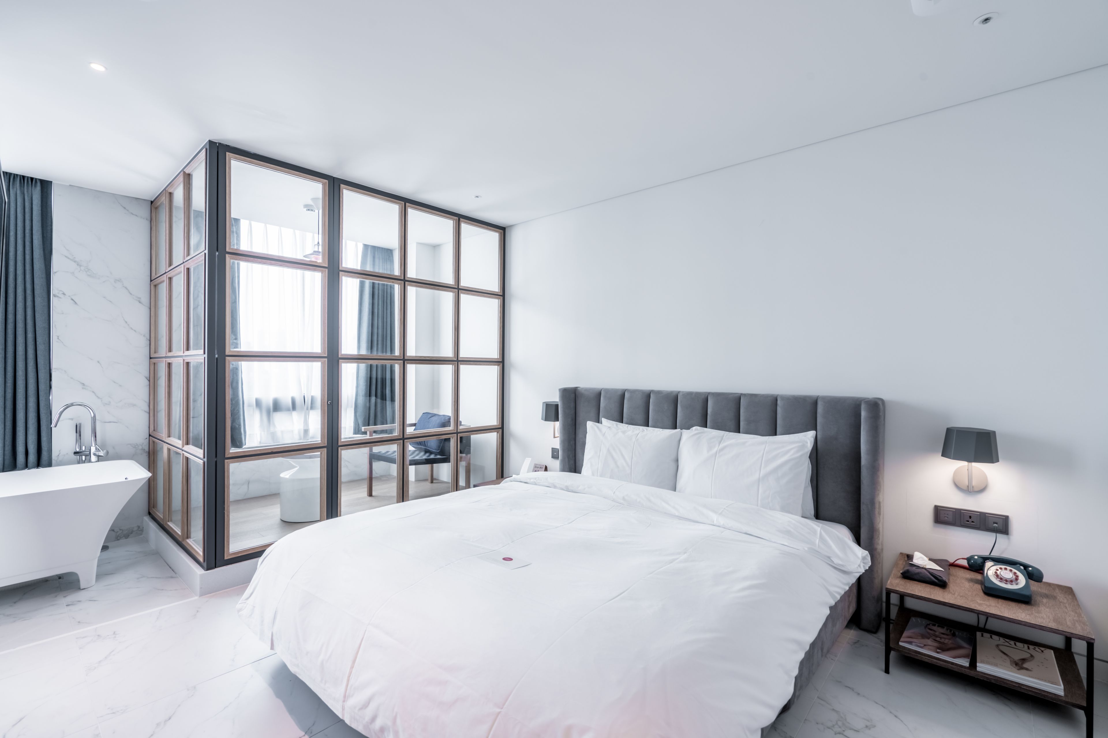 A modern bedroom with a large bed, a glass-enclosed bathroom, and a nightstand with a lamp.