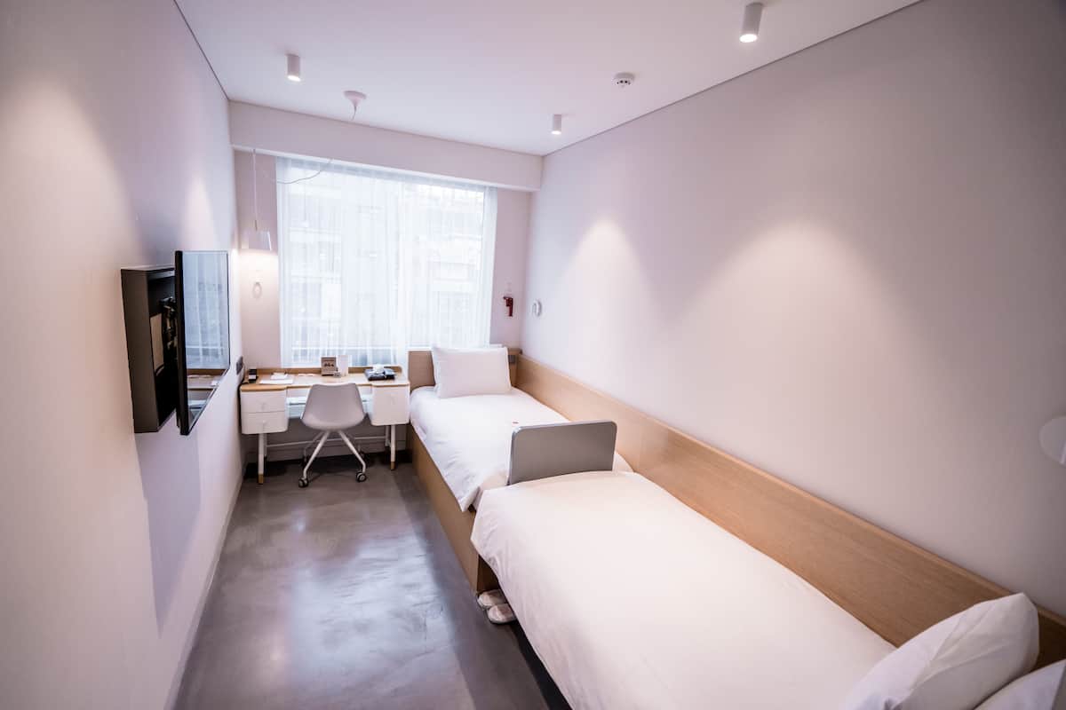 Una habitación de hotel moderna con dos camas individuales, un escritorio con silla, un televisor montado en la pared y un ventanal que proporciona luz natural.