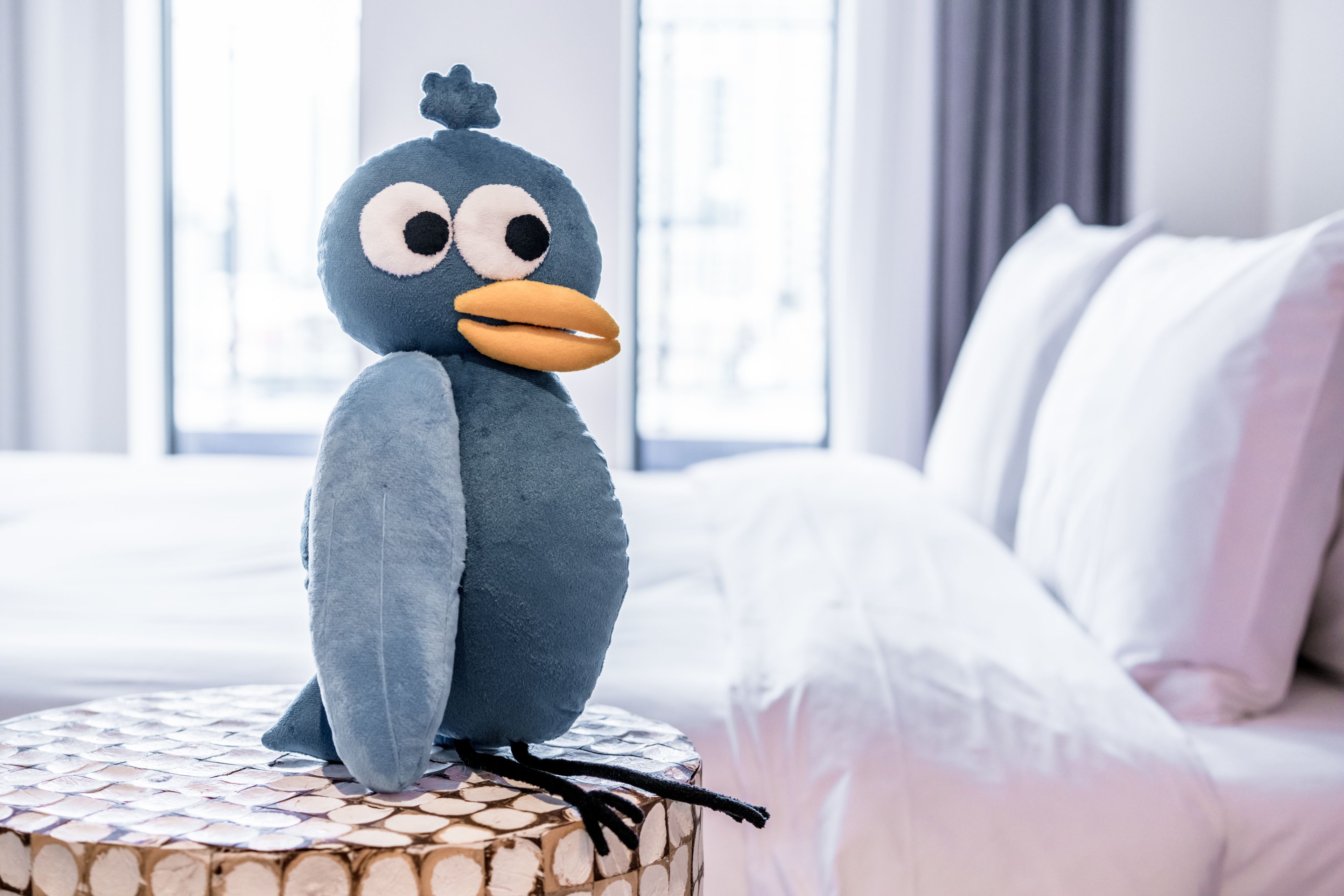 Un peluche sobre una mesa redonda en una habitación de hotel, con una cama y cortinas al fondo.