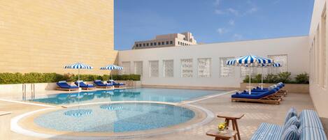 A swimming pool with lounge chairs and umbrellas, surrounded by a low wall and a building.