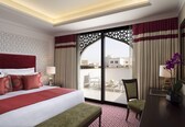 A hotel room with a large bed, a desk, a chair, and a balcony with a view of buildings.