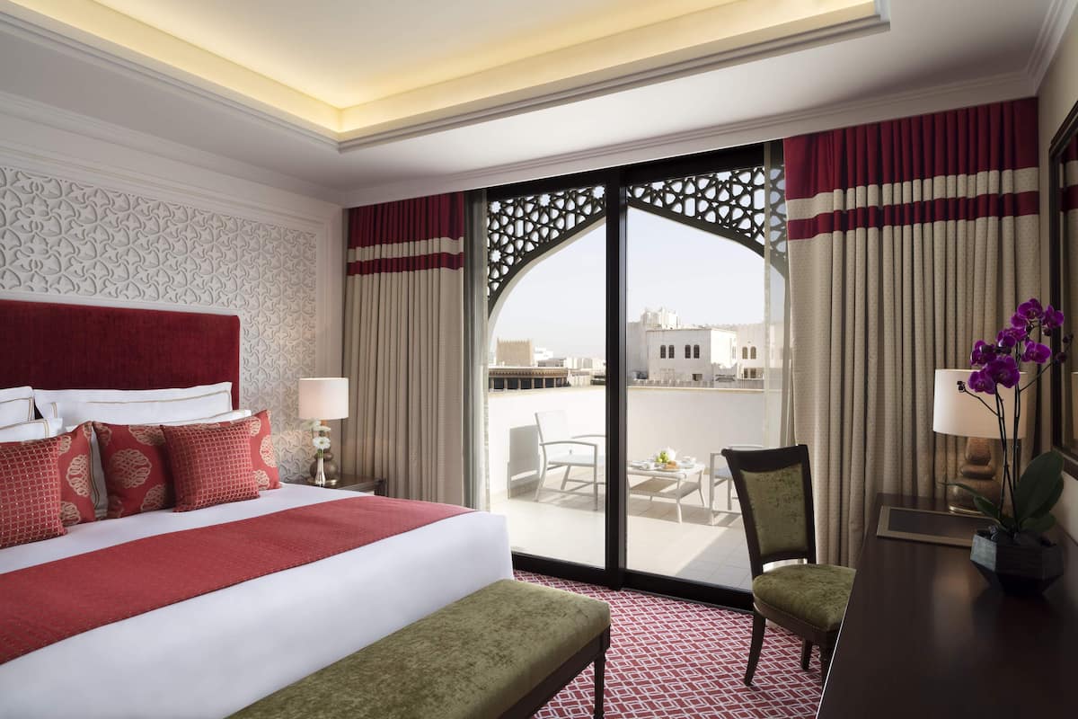 A hotel room with a large bed, a desk, a chair, and a balcony with a view of buildings.