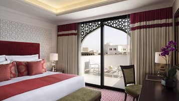 A hotel room with a large bed, a desk, a chair, and a balcony with a view of buildings.