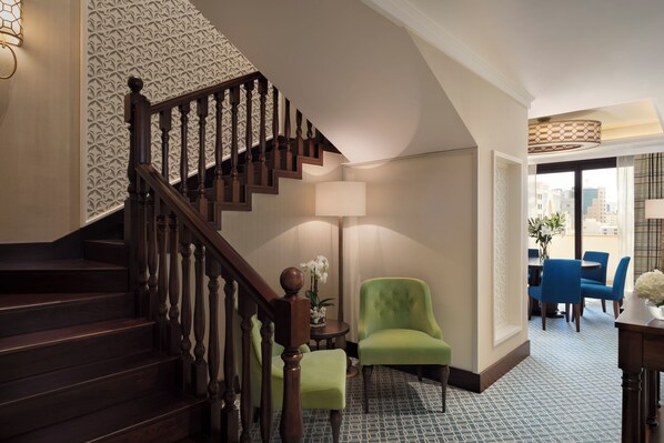 Una escalera con balaustres de madera, un sillón verde, una mesa redonda con una lámpara y un comedor con sillas azules.