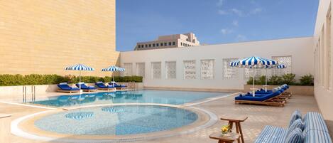 A rooftop swimming pool with lounge chairs and umbrellas, surrounded by a high wall and a building in the background.