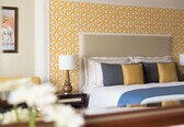 A hotel room with a patterned wallpaper, a bed with multiple pillows, a bedside table with a lamp, and a framed picture on the wall.