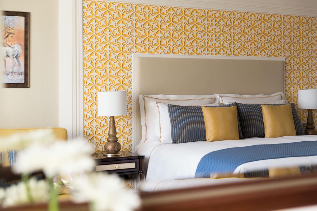 A hotel room with a patterned wallpaper, a bed with multiple pillows, a bedside table with a lamp, and a framed picture on the wall.