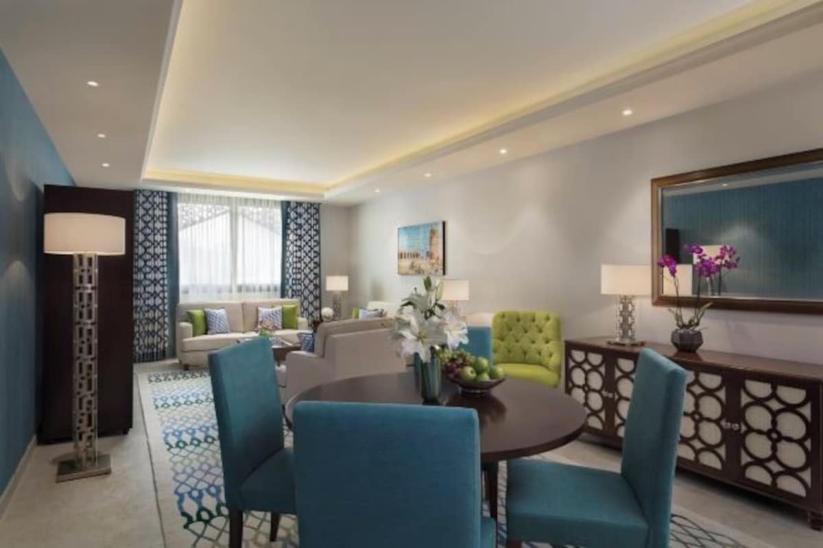 Una habitación de hotel moderna con zona de comedor, un sofá, un sillón verde y un espejo grande.