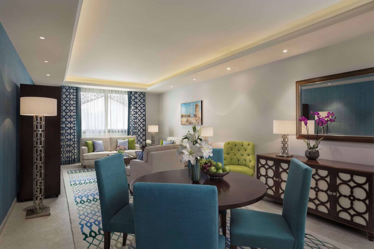 Een moderne hotelkamer met een zithoek, een bank, een groene fauteuil, een console tafel met spiegel en een groot raam met behang.