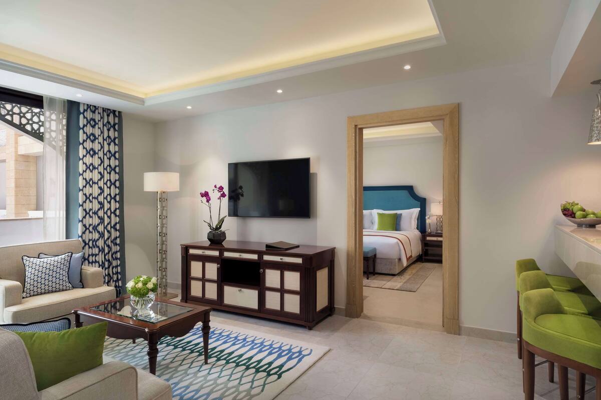 Een moderne hotelkamer met een sofa, een salontafel, een televisie, een bed en een eethoek met groene stoelen.