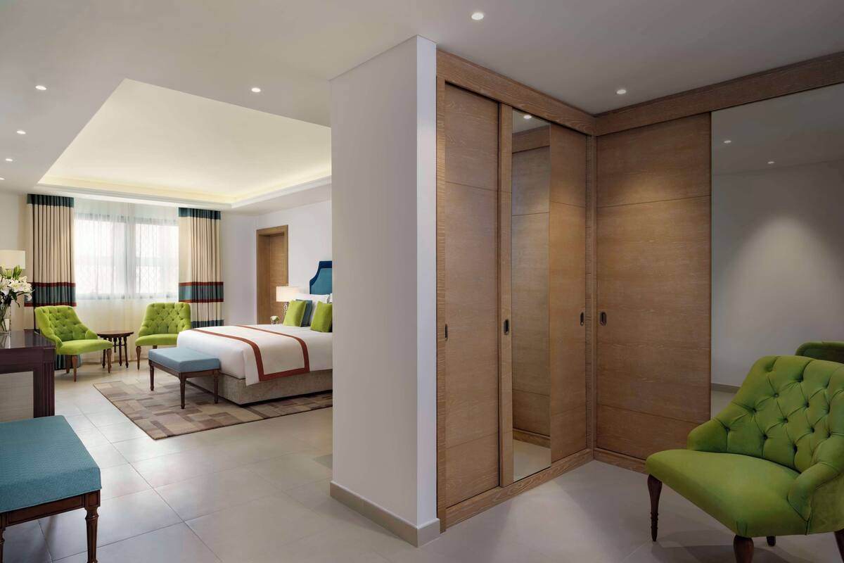 Een moderne hotelkamer met een bed, groene fauteuils, een klein tafeltje en een houten kledingkast.