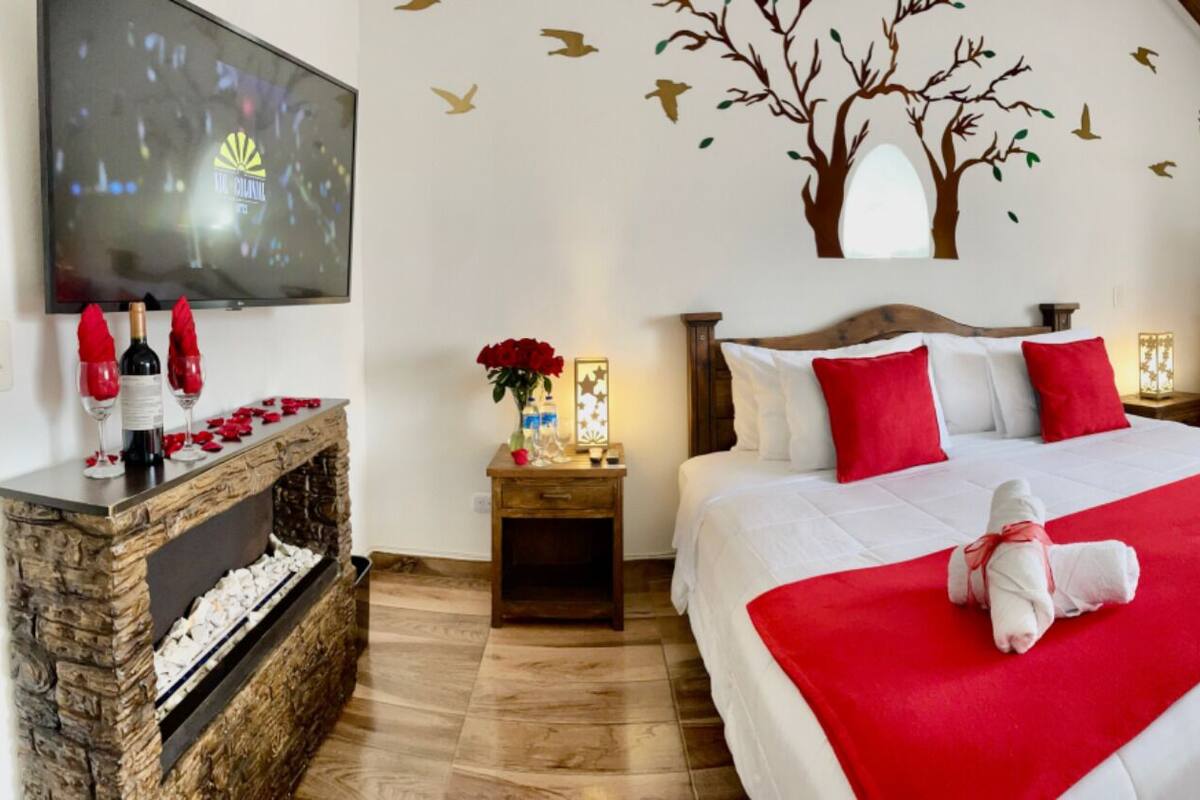 A hotel room with a large bed, a wooden headboard, a TV, a fireplace, a desk, and a bathroom with a bathtub.